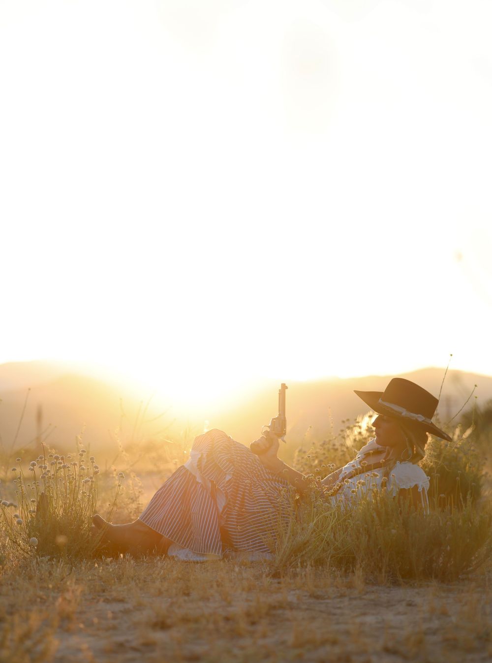 Two people sitting in golden field at sunset, one wearing a wide-brim hat, bathed in warm glowing light.
