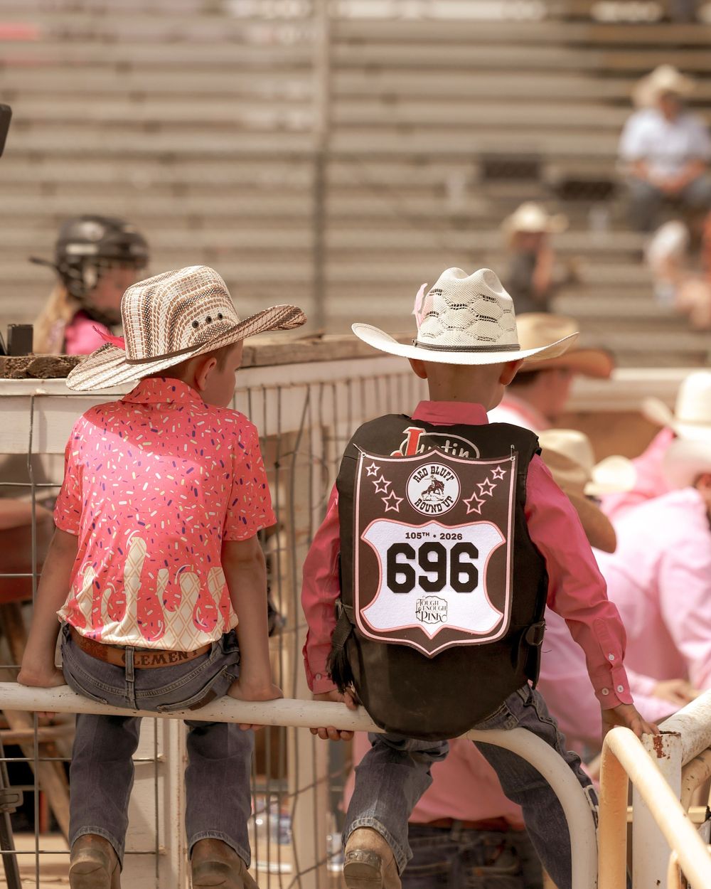 Two young cowboys in cowboy hats lean on a fence at a rodeo, one wearing a competition vest numbered 696.
