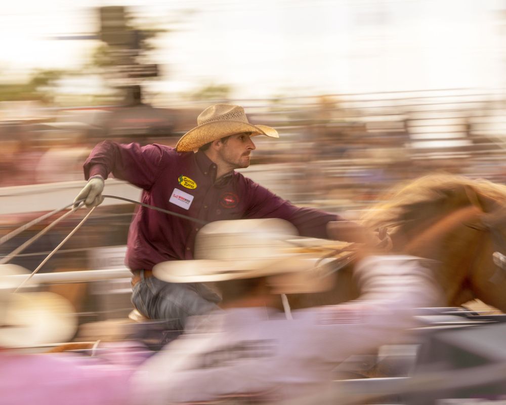 A cowboy in a hat leans over the arena fence during a fast-paced rodeo event.