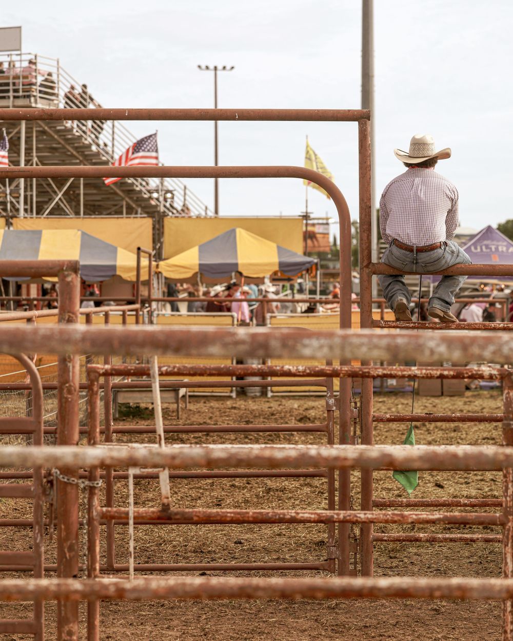 A cowboy sits atop a rusty metal gate overlooking a rodeo arena with American flag.