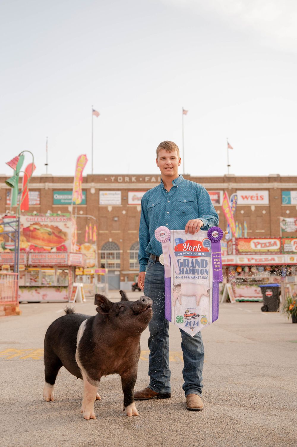 Person posing with a prize-winning pig at a county fair with carnival booths in background.
