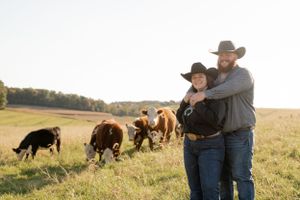 Couple embraces in pasture with herd of cattle grazing in background during sunset.