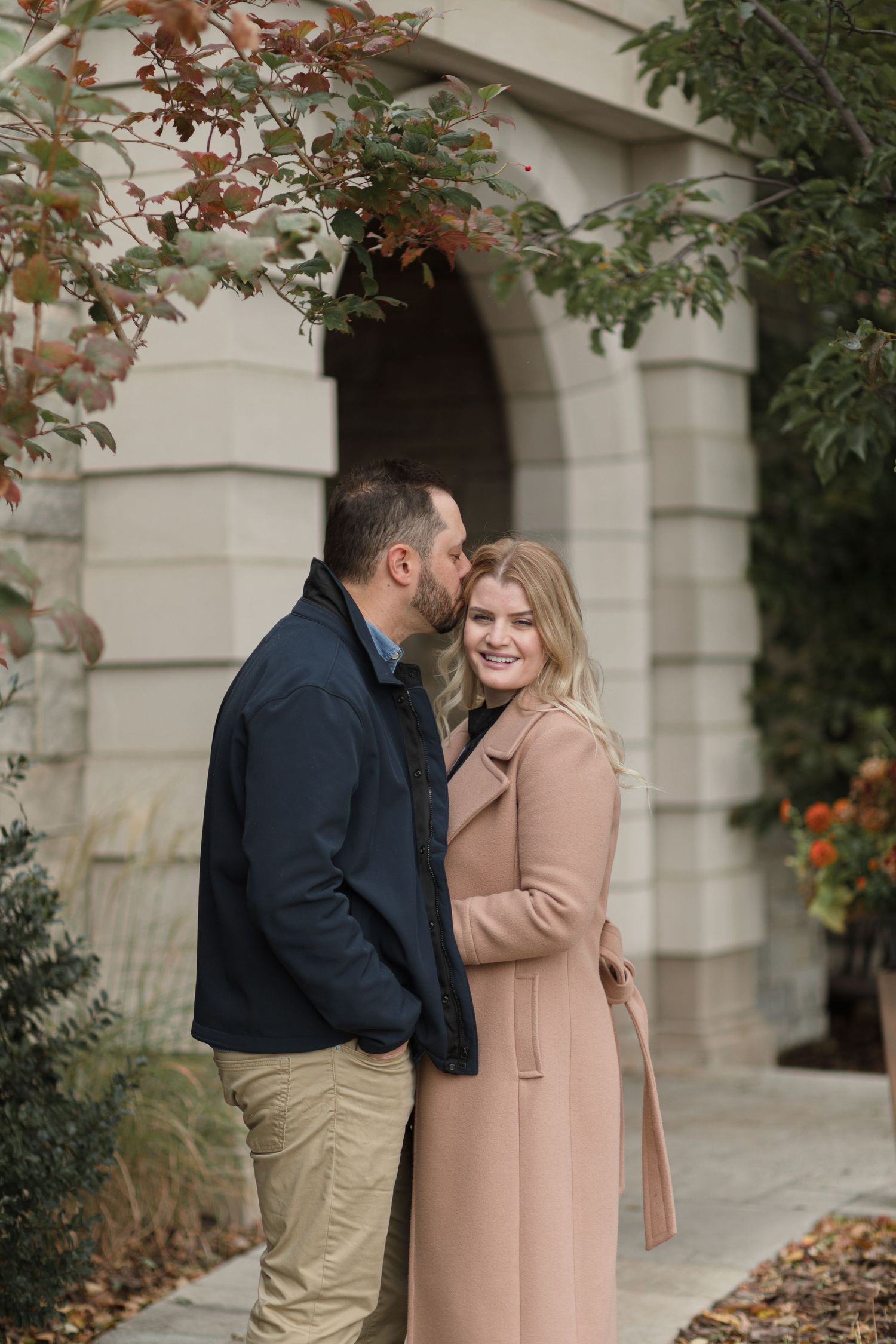 Couple sharing an intimate moment under stone archway surrounded by autumn foliage and orange flowers.