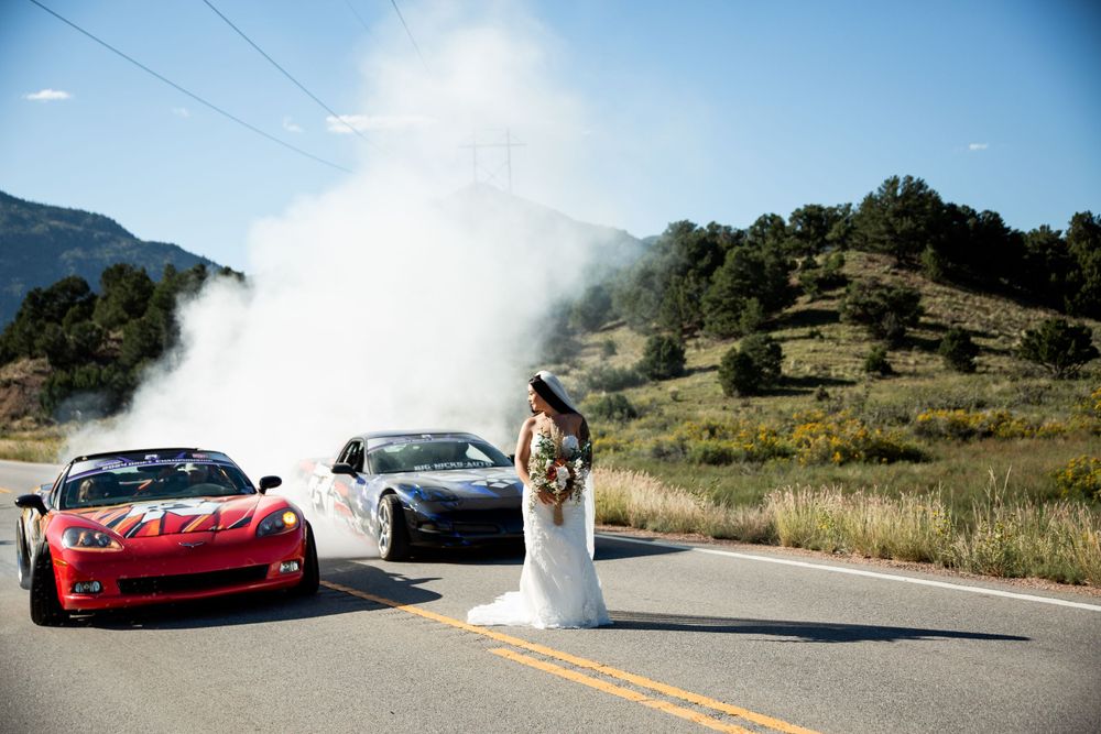 A bride poses between two sports cars on a mountain road with dramatic fog in the background.