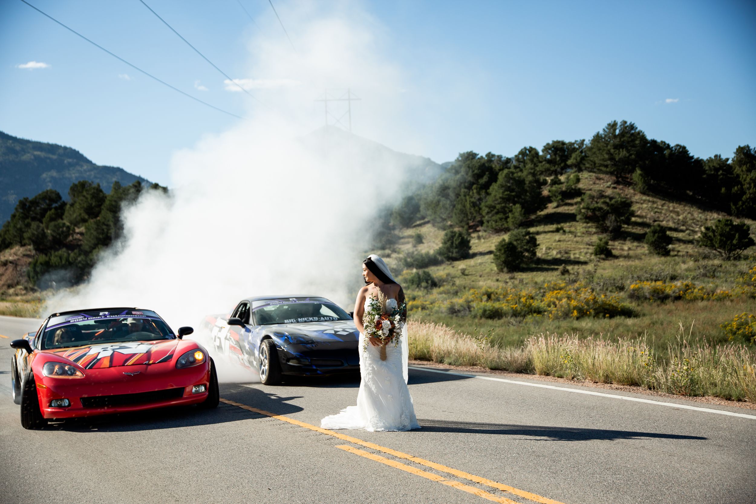 A bride poses between two sports cars on a mountain road with dramatic fog in the background.