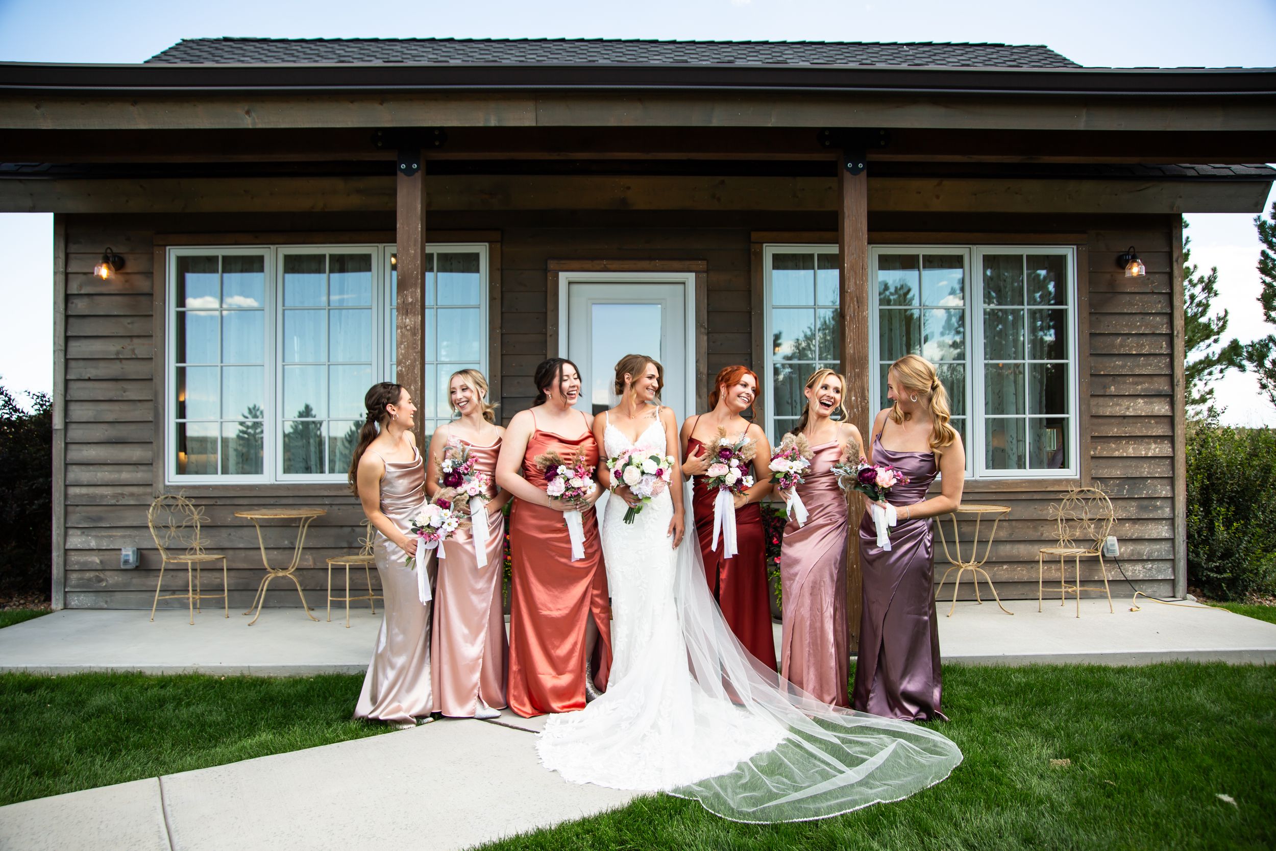 Bridesmaids in pink and mauve dresses gathered with bride outside rustic wedding venue.