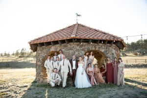 A wedding party poses together outside a rustic stone gazebo with wooden roof.