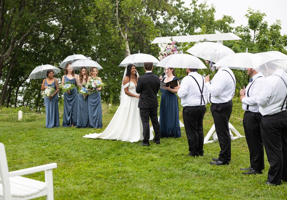 Wedding guests holding white umbrellas gather on a lush green lawn during an outdoor ceremony in the rain.