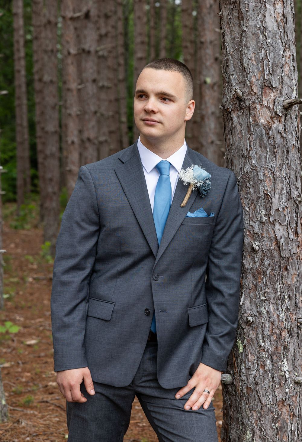 A groom in a grey suit with blue tie poses against pine trees in a forest setting.
