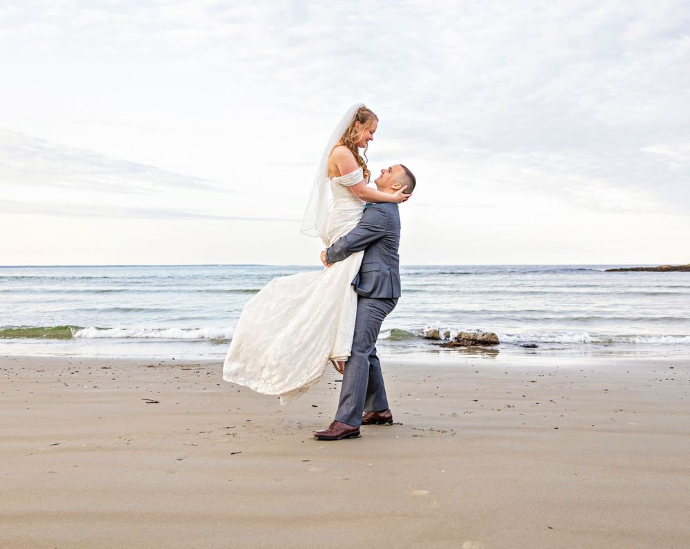 Romantic couple in wedding attire dance and embrace on a serene beach with waves and rocks in the background.