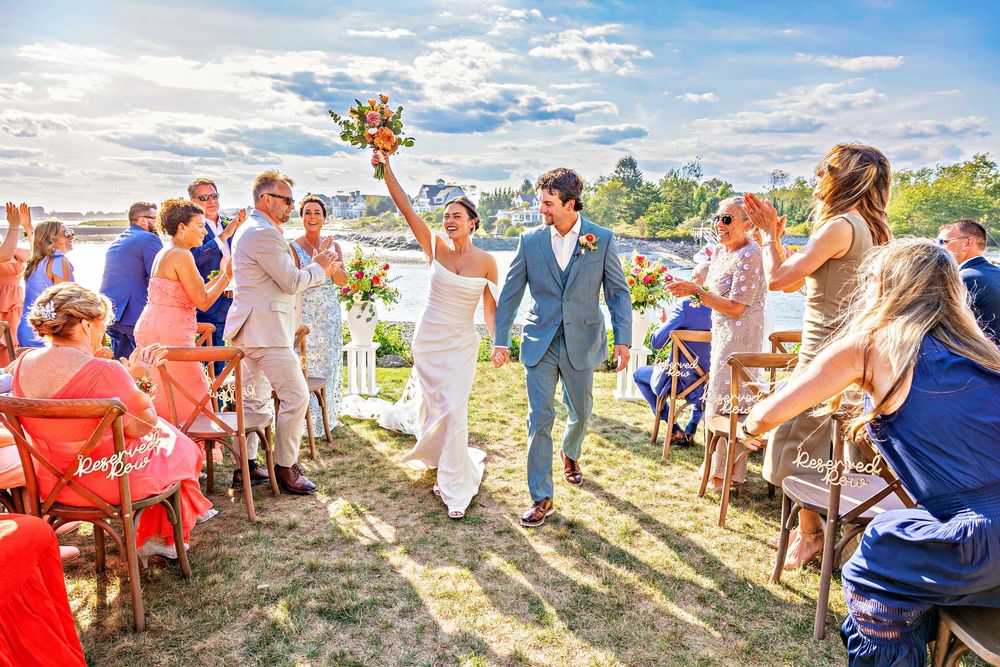 Wedding guests gather outdoors on a sunny hillside for a celebration.