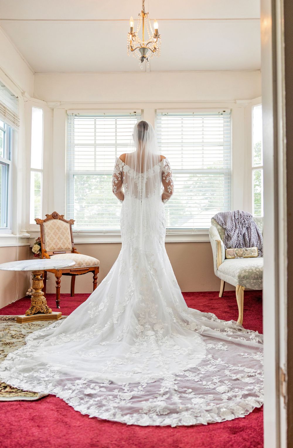 A stunning white lace wedding dress with long train displayed on red carpet in a bright sunlit room.
