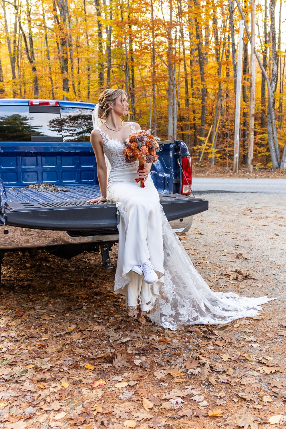 A wedding party poses in a beautiful autumn forest setting with golden leaves and a blue pickup truck in the background.