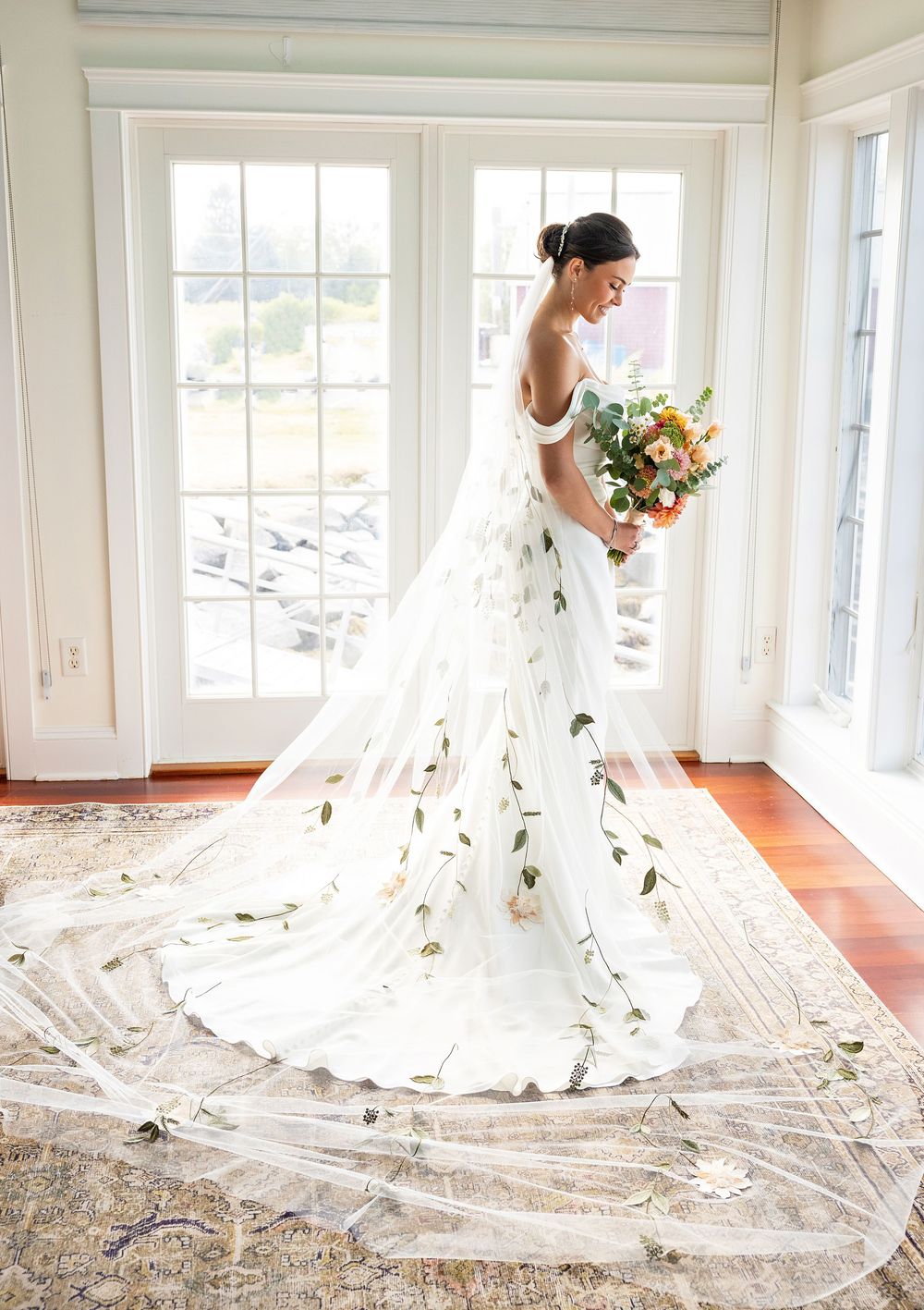 Elegant wedding dress with flowing cathedral veil adorned with delicate floral details stands by bright bay windows.