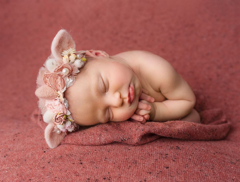 Peaceful newborn sleeping on soft rose-colored fabric wearing delicate floral headband.