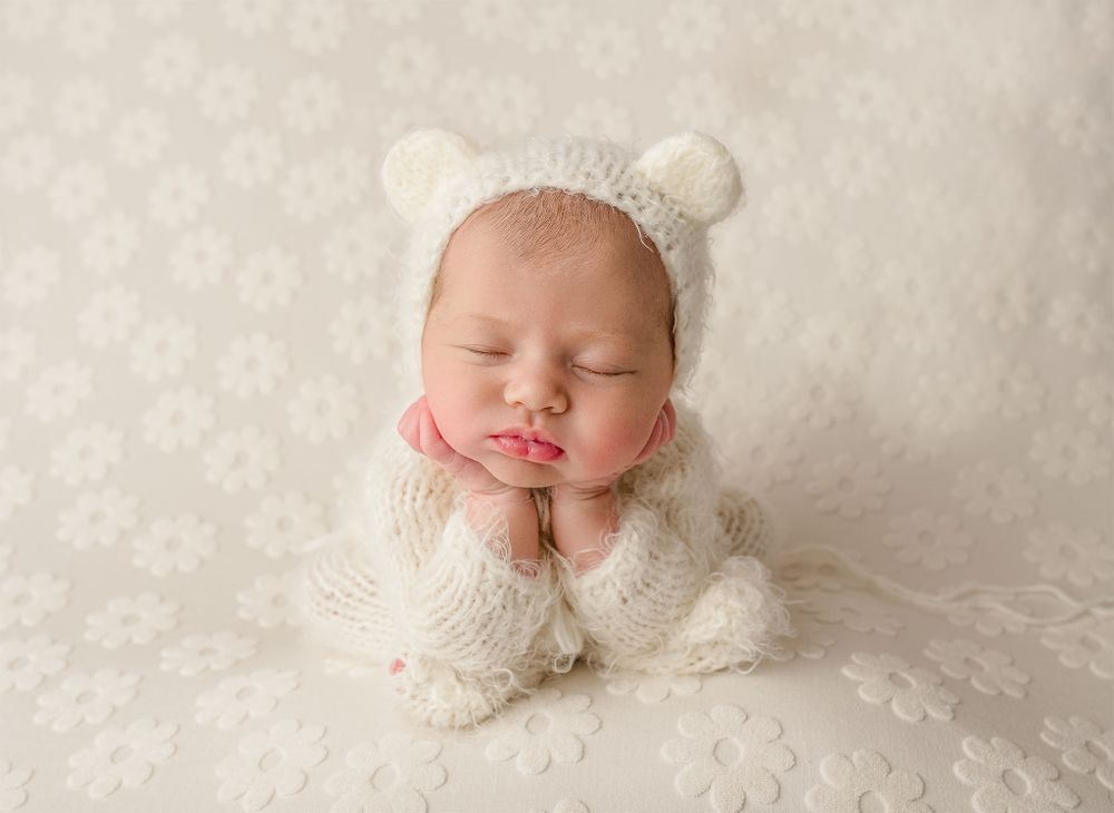 Sleeping newborn in a white knit bear-ear bonnet and outfit lays on a textured white backdrop during a studio session.