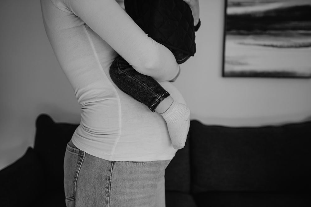 Black and white photo of a pregnant woman in jeans cradling her baby bump while holding a dark jacket indoors.