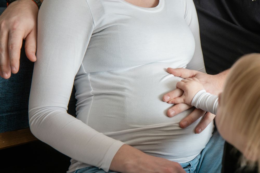 A child's hands gently touching a pregnant woman's baby bump through a white long-sleeve shirt.