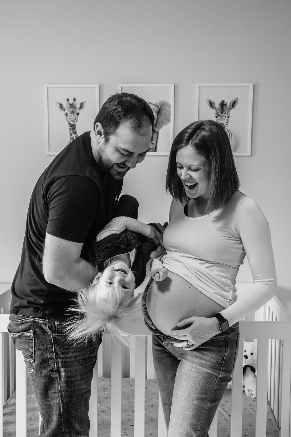Black and white photo of laughing expectant parents holding toddler upside down in nursery with animal wall art.