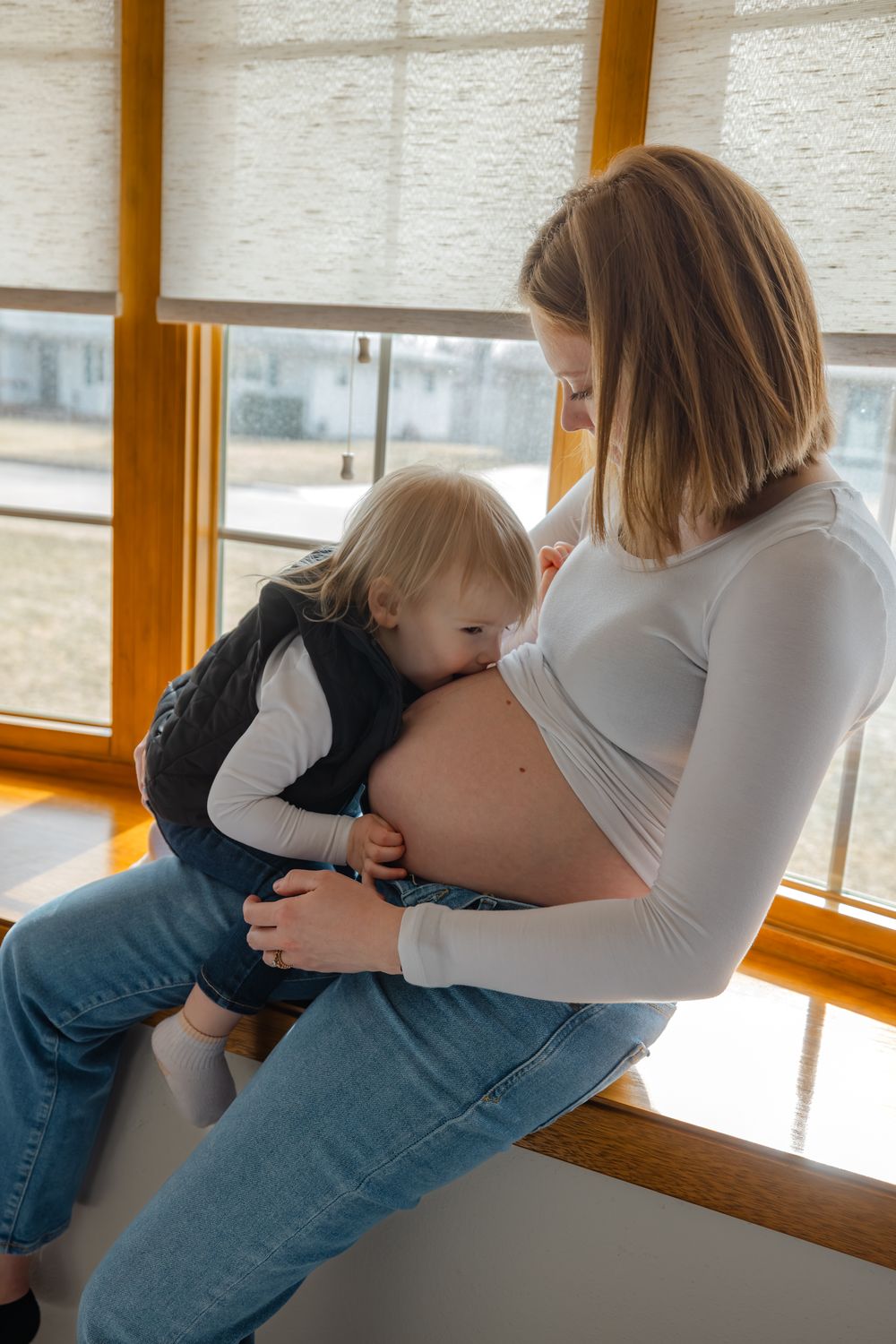 Pregnant woman in white top and jeans holding toddler child near window, child resting head on baby bump.