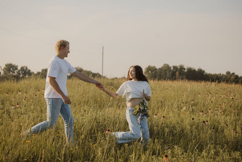Couple in casual attire playfully running through a golden grassy meadow at sunset.