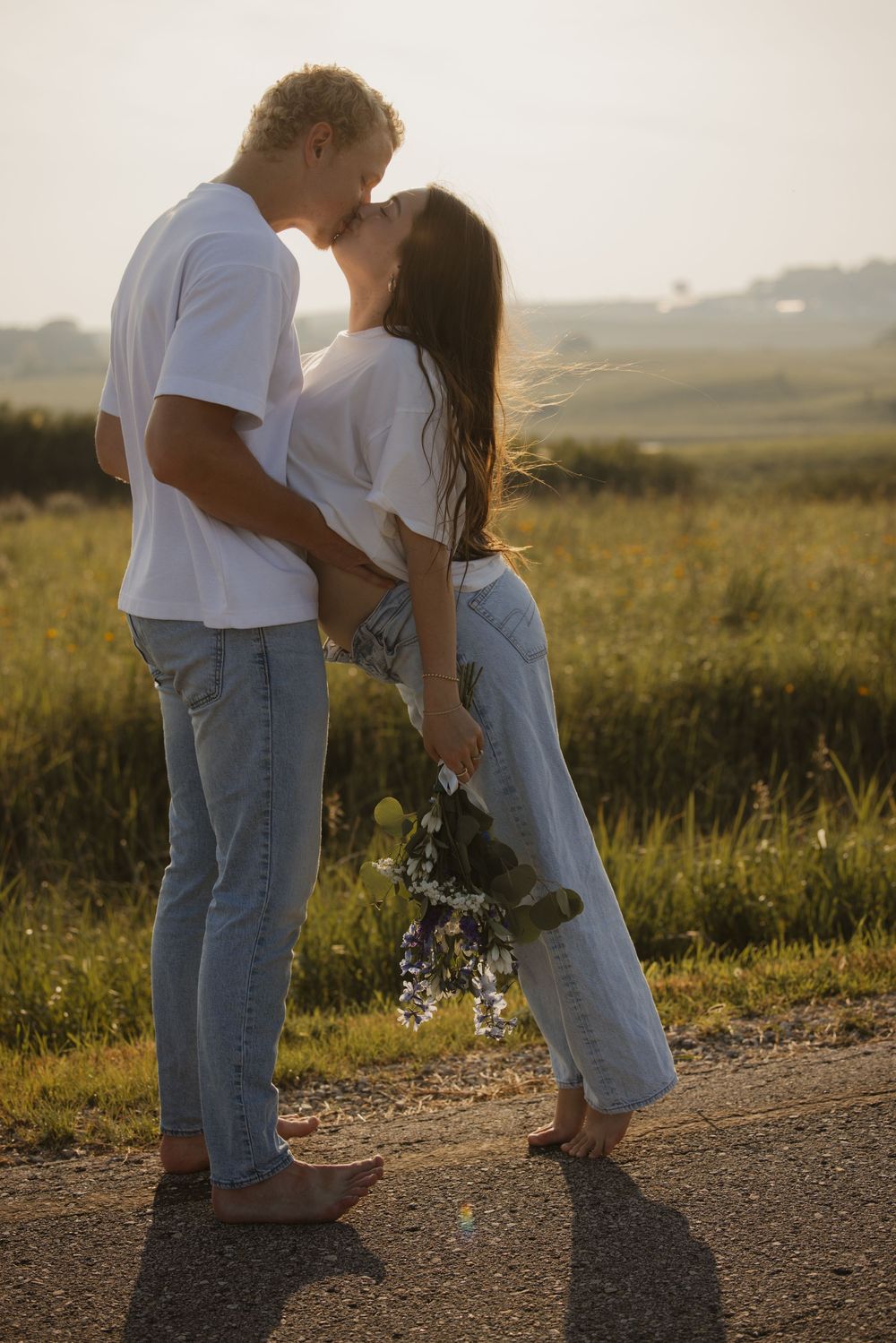 A couple in white shirts and jeans share a tender moment together in a golden field at sunset.