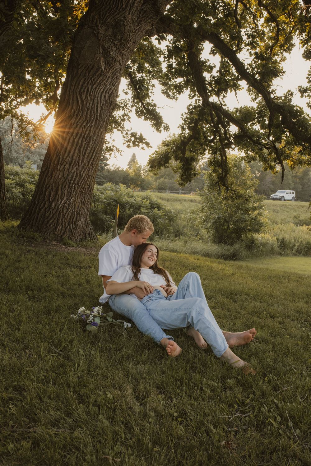 A couple sits together under a large oak tree at sunset in a grassy meadow.