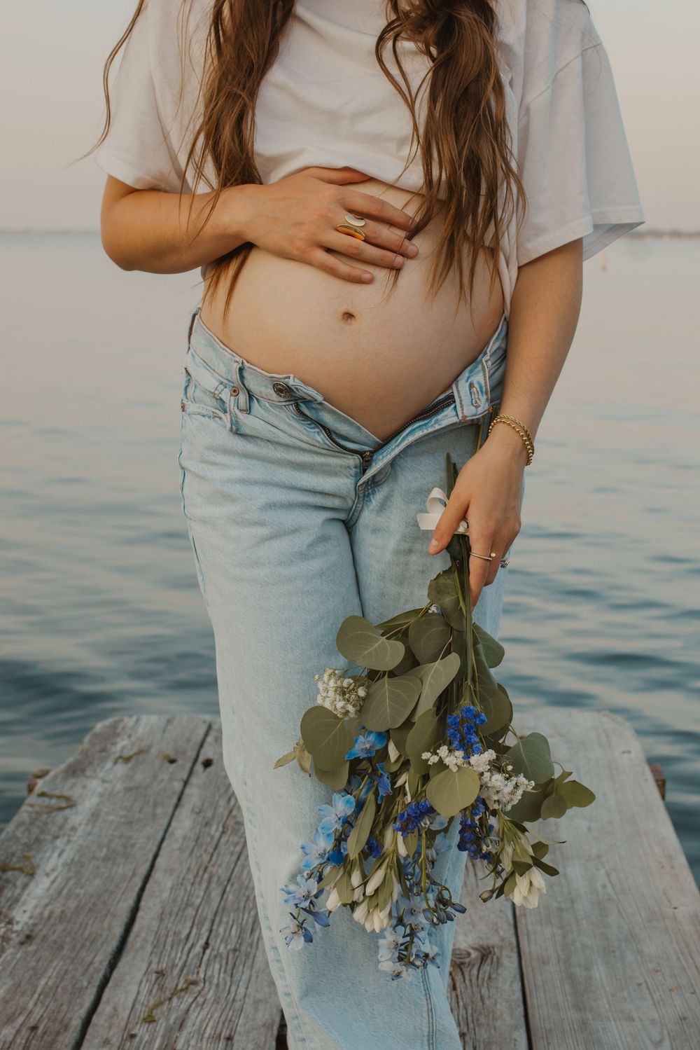 Person in light jeans holds blue and white flowers while standing on wooden dock by calm water.