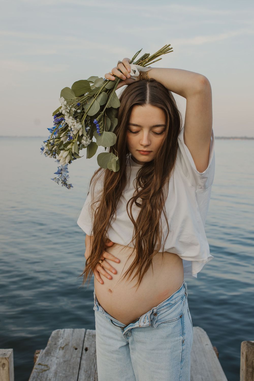 Woman in white top and jeans poses gracefully with flowers by serene waterfront in artistic maternity photoshoot.