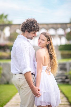 Romantic couple in white outfits holding hands in a garden, woman smiling over shoulder, arched architecture in background.
