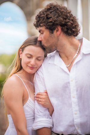 A couple in white outfits share a tender moment, man kissing woman's temple while she smiles softly.