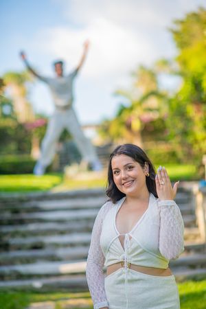 Smiling woman in white outfit shows engagement ring in park, with man jumping joyfully on steps behind her.