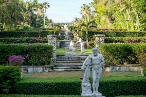 Classical stone statues and manicured hedges adorn a formal garden with stone steps and tropical palms.