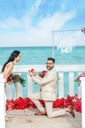 Man proposing on one knee with ring beside ocean, red roses and Will You Marry Me sign backdrop.