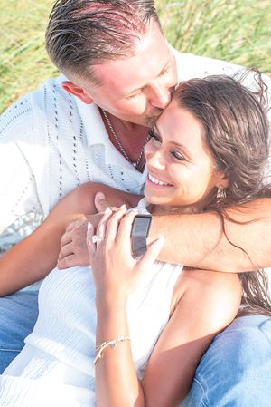 Couple sitting outdoors, man kissing woman on head while she smiles, holding hands near grassy field.