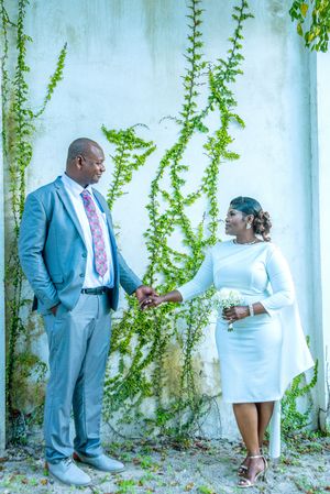Couple in wedding attire holding hands before ivy-covered wall, man in gray suit, woman in white dress holding flowers.