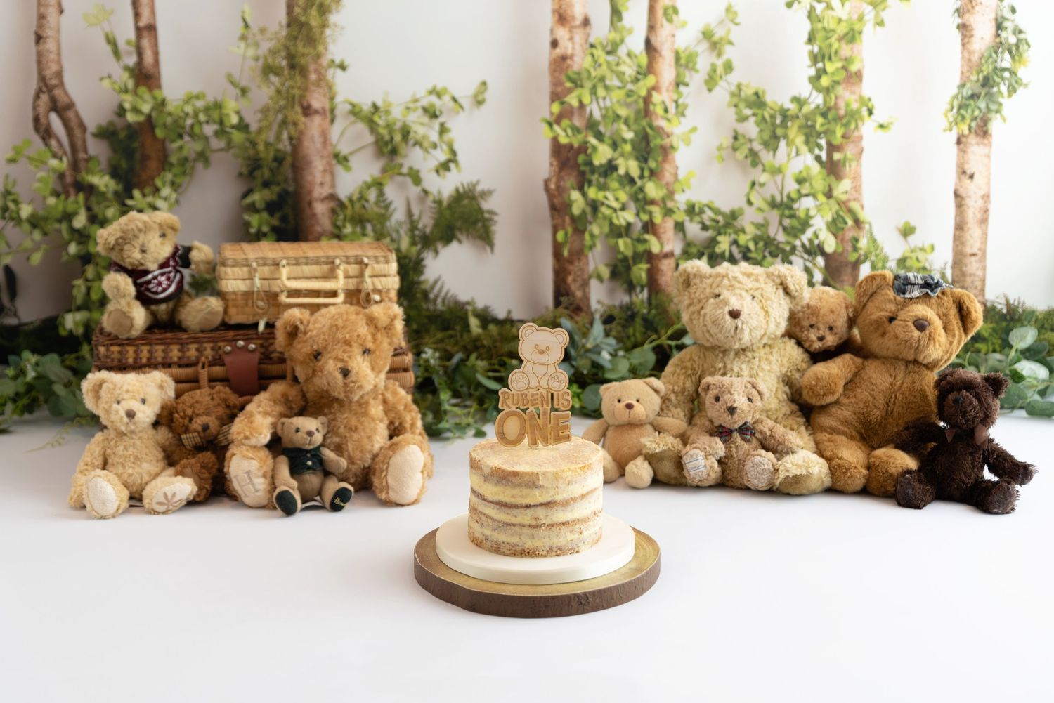 A group of teddy bears arranged around a tiered gold and white cake against a white backdrop with greenery.