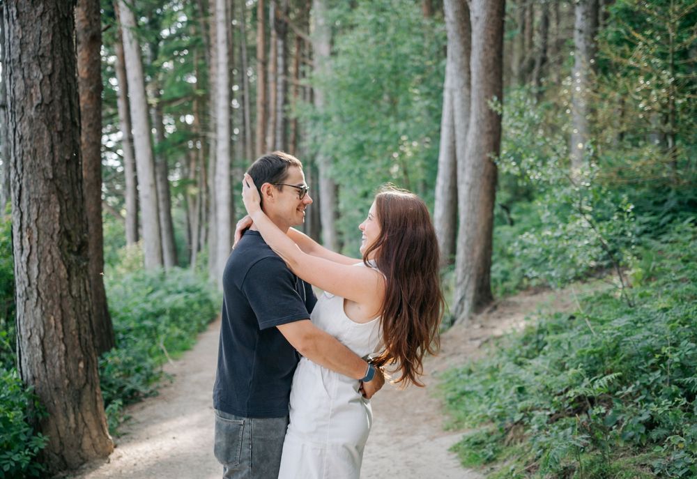 A loving couple embraces on a forest path surrounded by tall evergreen trees and lush greenery.