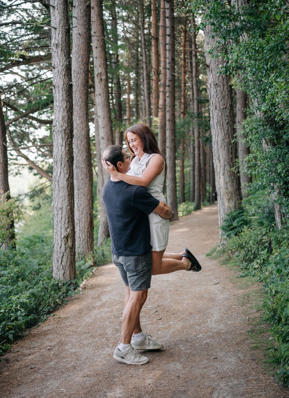 A couple embraces on a scenic forest trail lined with tall pine trees on a sunny day.