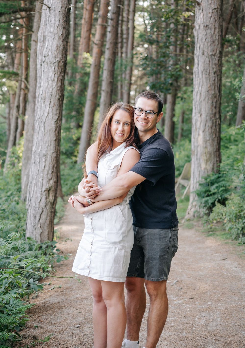 Two people embrace and share a joyful moment while standing along a serene forest path lined with tall pine trees.