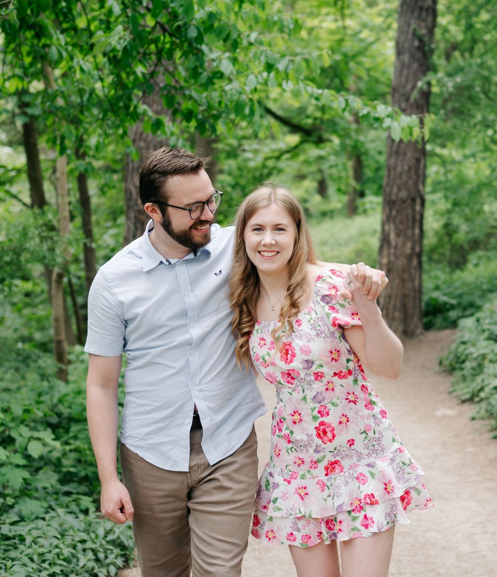 A couple walking together on a wooded path, with lush greenery surrounding them on a sunny day.