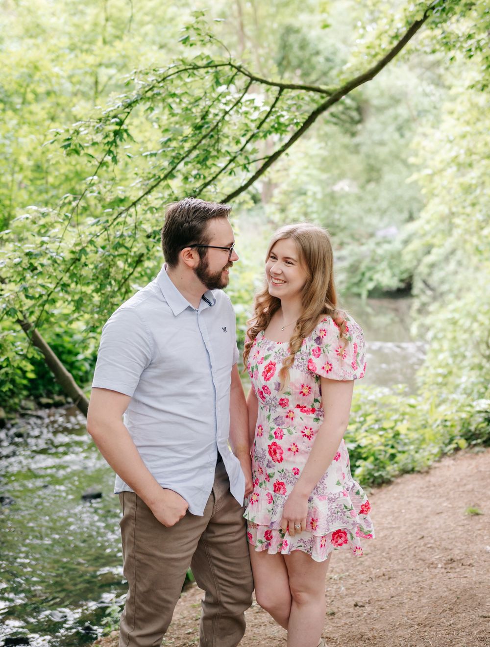 A couple stands together on a dirt path by a stream, surrounded by lush green trees and foliage in a peaceful outdoor setting.