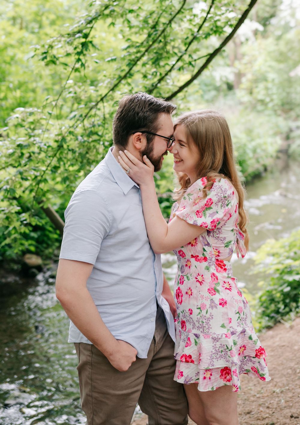 A couple shares a tender moment on a nature trail surrounded by lush greenery beside a stream in a romantic setting.