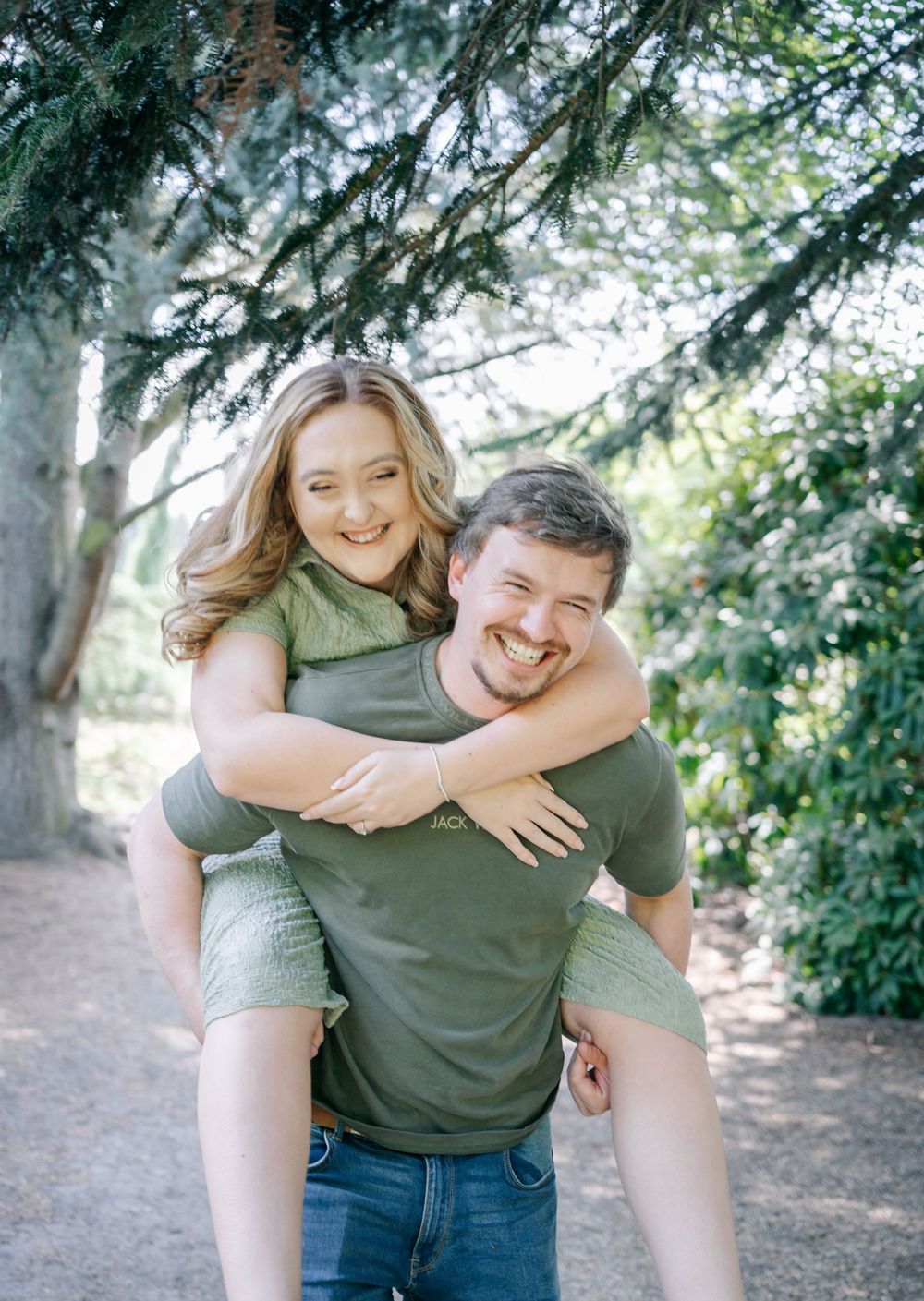 A couple shares a joyful piggyback ride moment on a trail surrounded by evergreen trees and natural scenery.