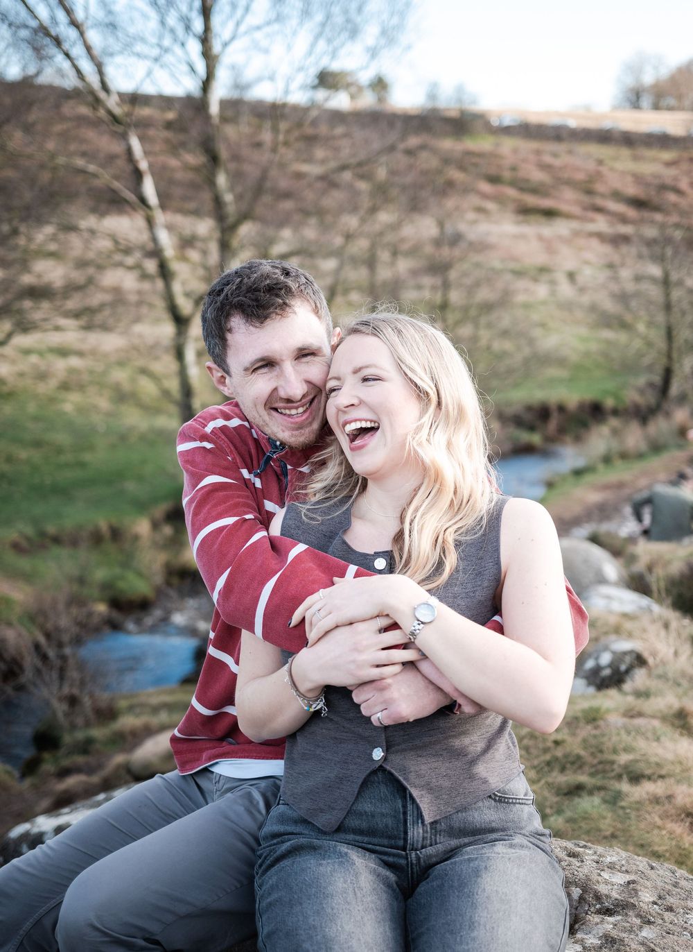 Couple sharing joyful laughter while sitting together on rocks near a stream on a spring day.