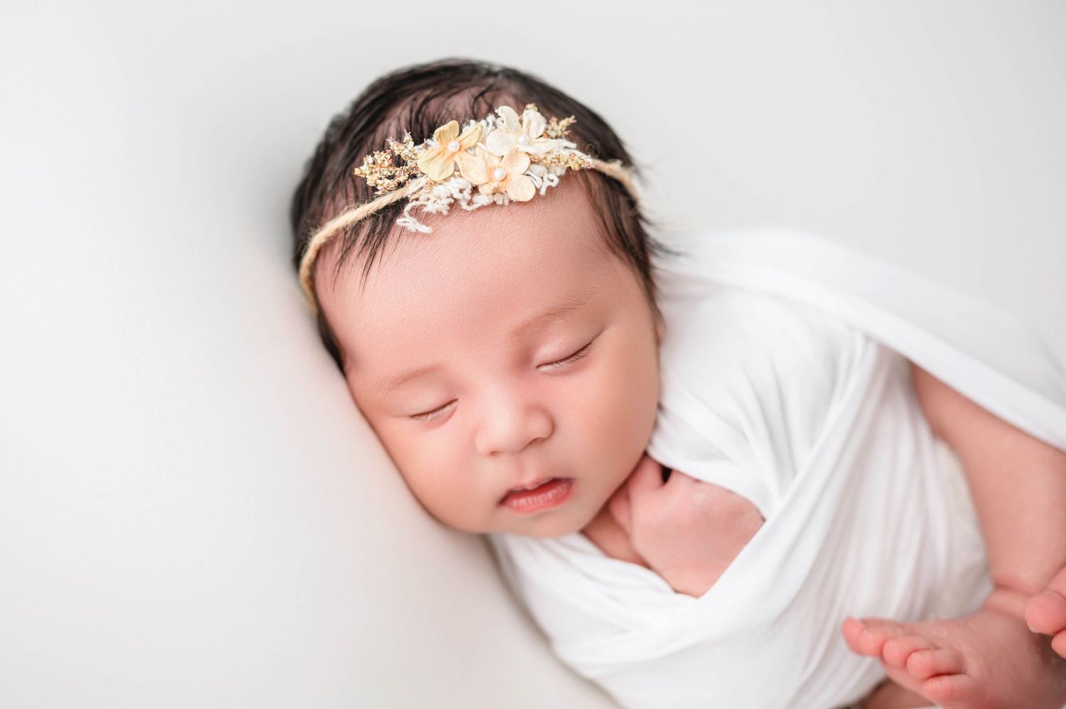 Baby girl in a floral headband, resting on a soft white blanket – timeless newborn portrait
