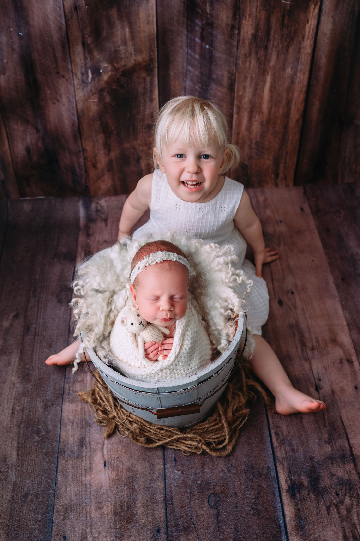 Newborn baby resting peacefully in a rustic bucket, with older sibling gently leaning over and smiling at the baby