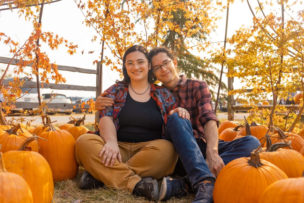 A couple sitting on the ground surrounded by pumpkins leaning on each other.
