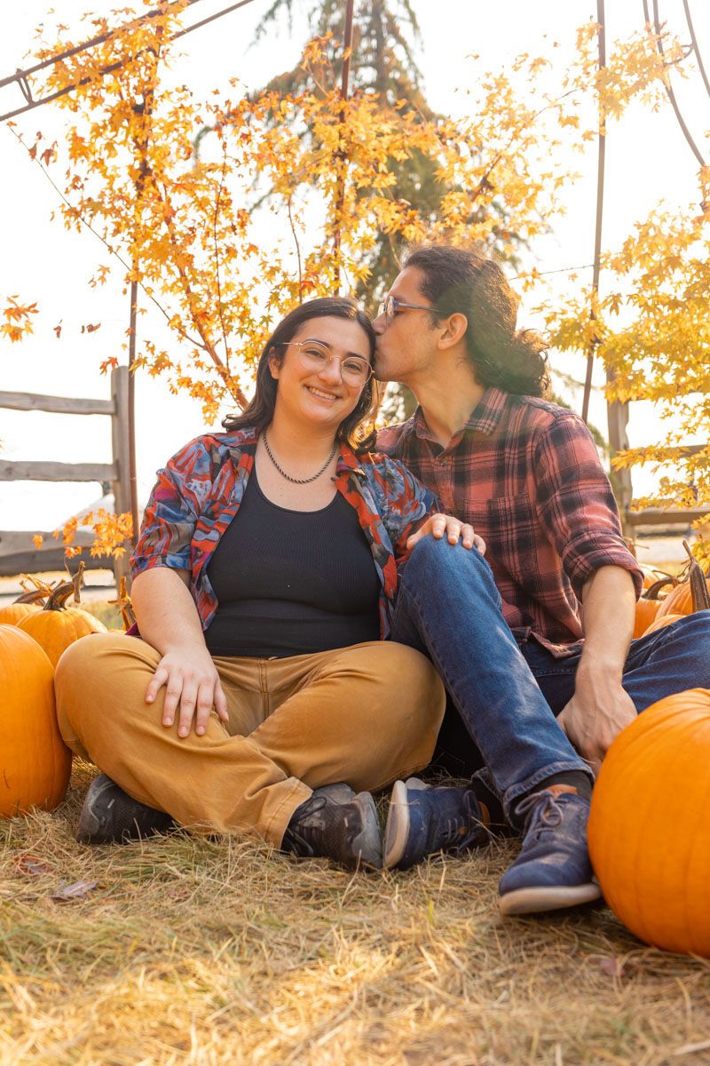 A couple sitting on the ground surrounded by pumpkins with one person kissing the others head.