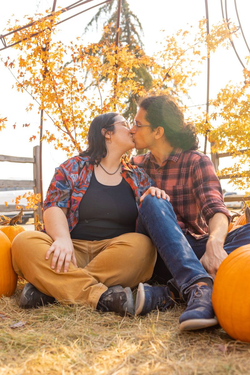 A couple sitting on the ground surrounded by pumpkins kissing.